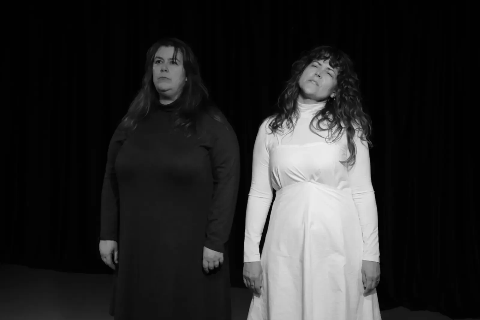 Two performers stand side by side against a black curtain, one dressed in black, one in white